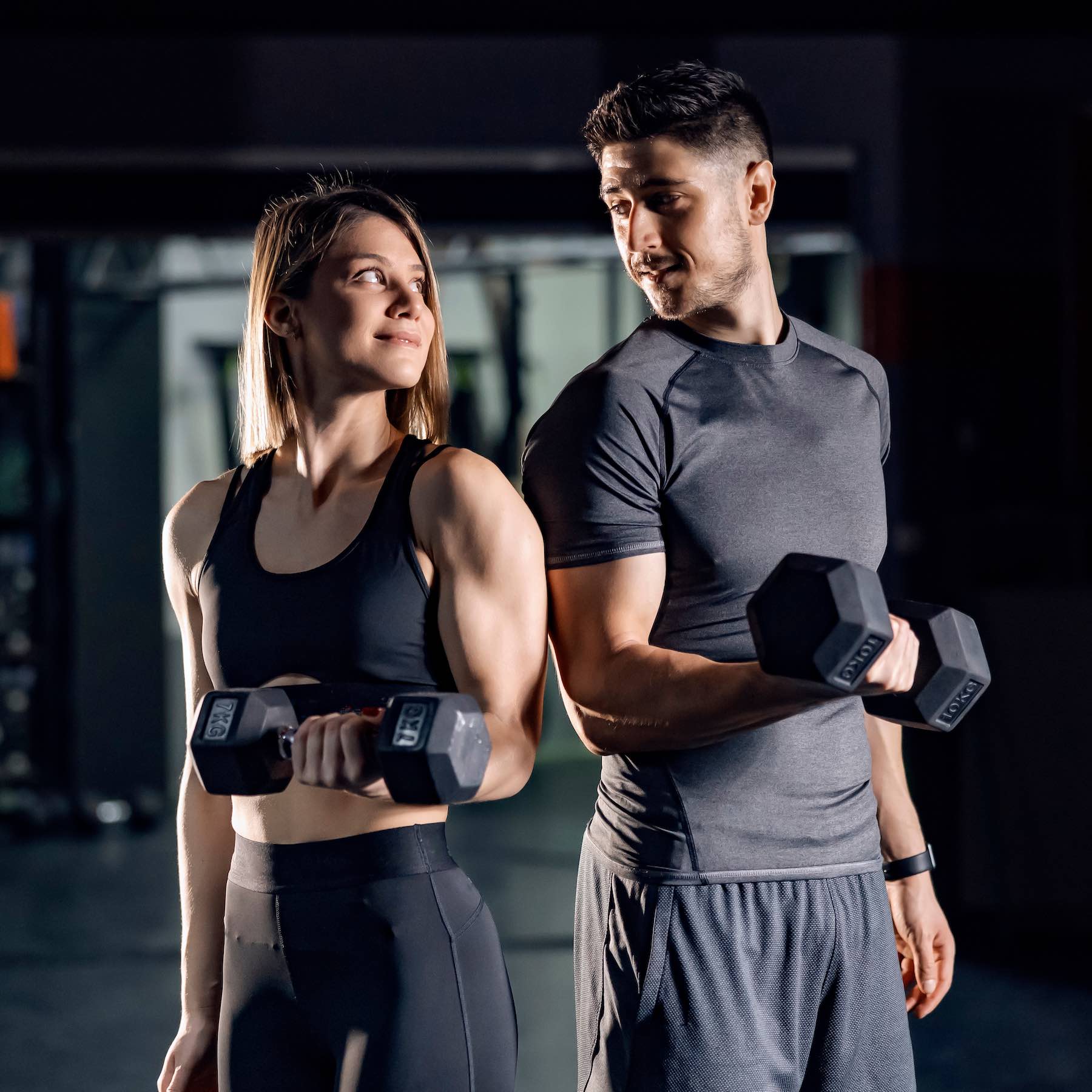 A happy sporty couple doing exercises for biceps with barbells in a gym and making eye contact with each other.